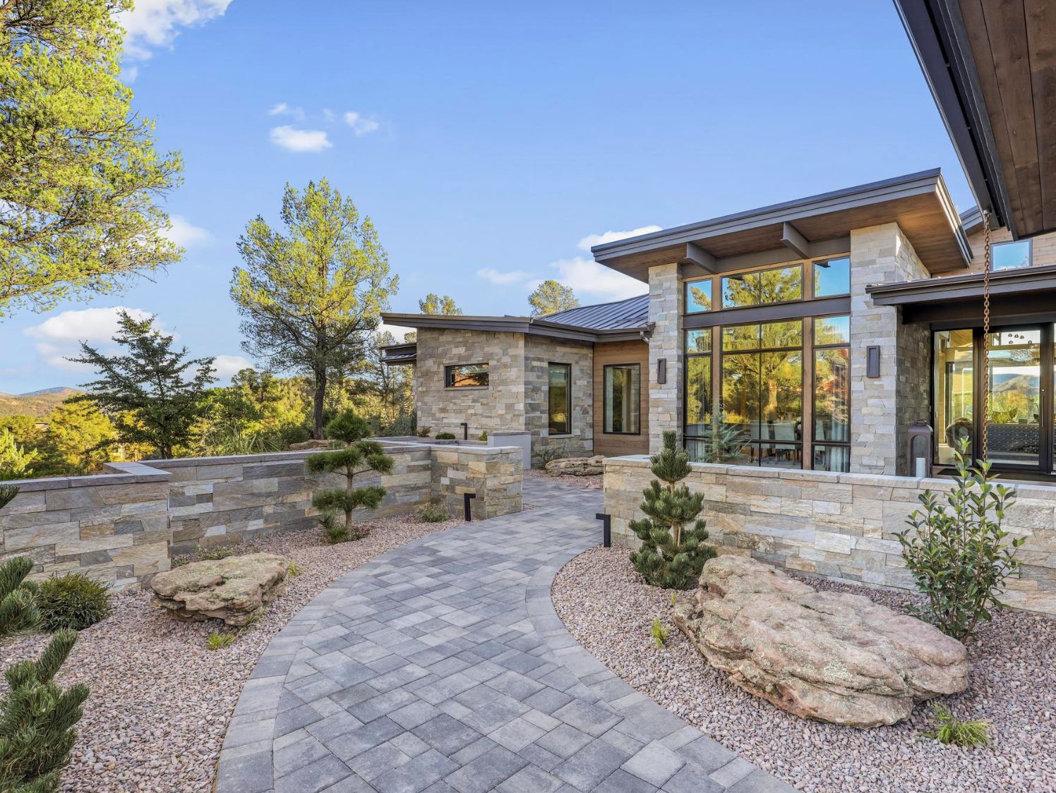 Modern stone house with large windows and a curved paved walkway surrounded by landscaping rocks and small trees.